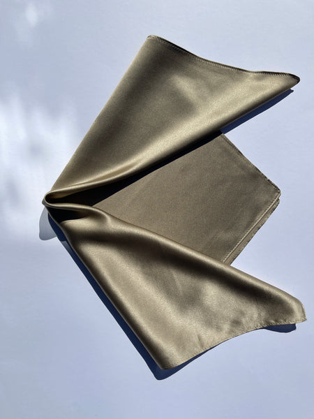 A folded silk square scarf in a muted olive-gold or khaki-tan color, displayed against a light blue-gray background.