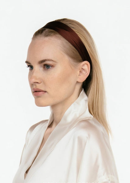 A blonde woman in profile wearing a dark reddish-brown wide silk headband and a cream satin shirt, against a plain white background. 