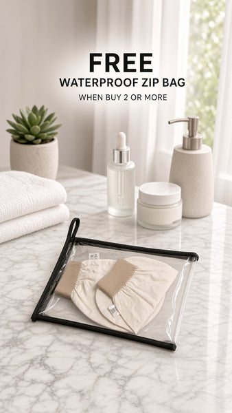 A clear waterproof zip bag with a black frame and black zipper pull is shown on a marble countertop, containing a beige fabric pouch. Folded white towels, a small potted succulent, skincare bottles, and a soap dispenser are arranged in the background near a window.
