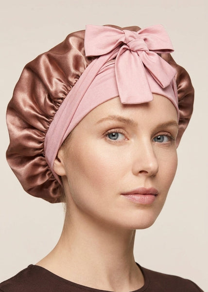 A woman wearing a silk sleep cap in two colors: a dusty pink headband with a large bow and adjustable elastic ties She is looking directly at the camera against a plain light background.